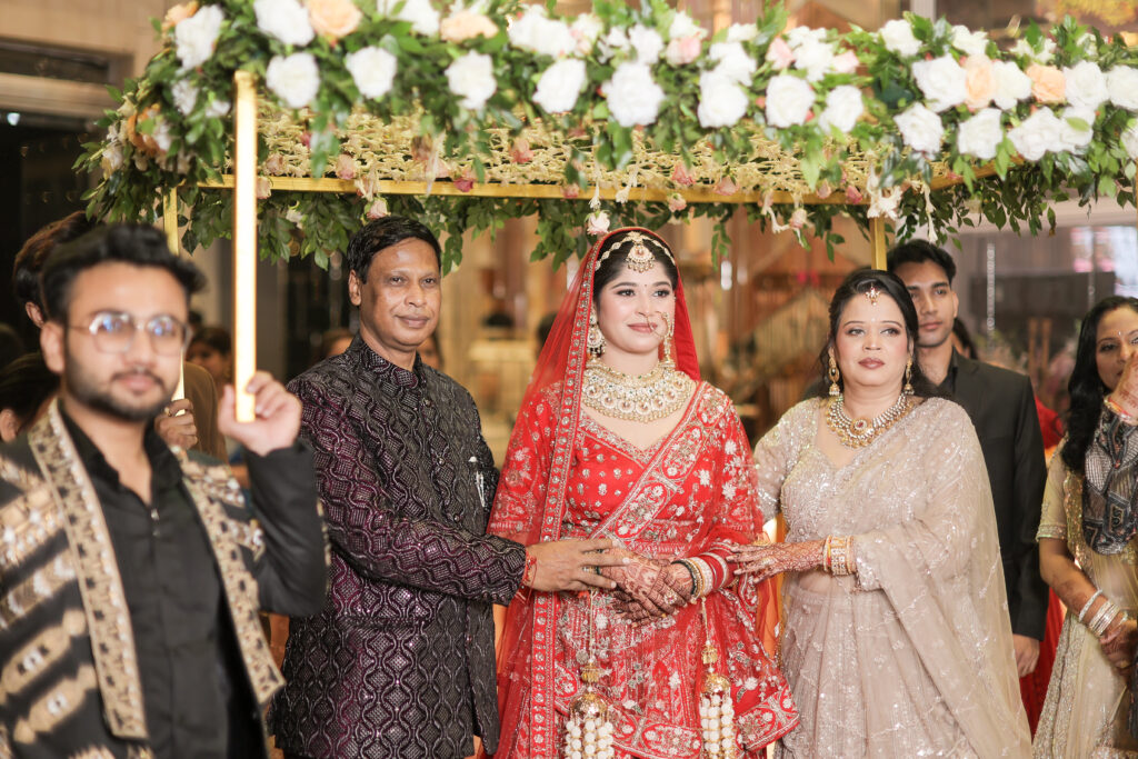 An indoor group portrait of a bride in a radiant red lehenga and veil standing beneath a decorated floral canopy (phoolon ki chaadar) held by family members. She is flanked by her parents, with guests and family members surrounding her during the grand wedding entry.