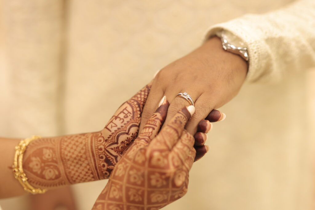 A sophisticated macro photograph capturing a detailed engagement ritual. A hand adorned with intricate mehendi places a refined diamond solitaire ring onto a partner's finger, highlighting craftsmanship, tradition, and subtle textures.