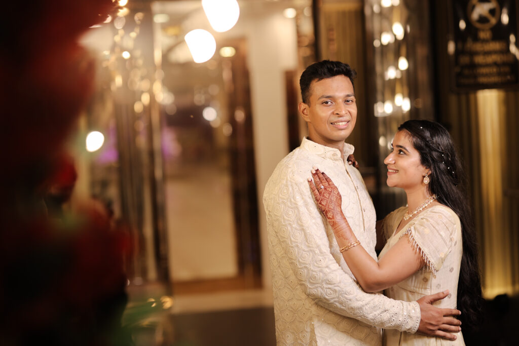 An indoor portrait of a couple embracing softly in a warmly lit venue hallway. The man smiles toward the camera wearing an embroidered cream-colored kurta, while the woman looks up at him lovingly with mehendi on her hand, wearing a matching ivory outfit and subtle jewelry.