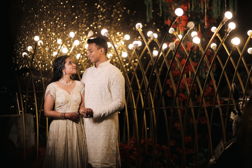 An outdoor portrait of a couple standing hand-in-hand beneath a decorative archway illuminated by dozens of glowing bulb lights and golden bokeh. The man wears an embroidered cream-colored kurta, and the woman wears a matching ivory gown with delicate jewelry and mehendi, looking up at him with a loving smile.