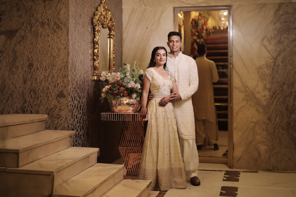 An indoor portrait of a couple posing together in a luxury hallway next to a marble staircase and a gold-framed mirror. A modern geometric table holds a vase filled with lush floral arrangements. The woman wears an embroidered ivory lehenga with silver detailing, and the man stands behind her wearing a matching white embroidered kurta and churidar set.