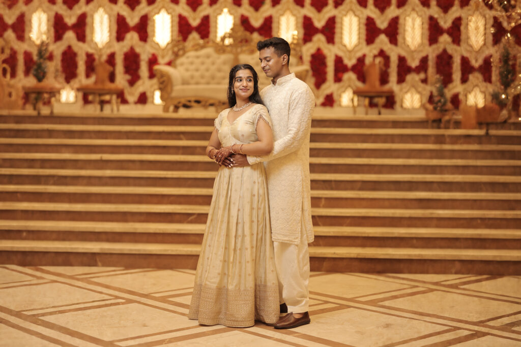 A wide indoor portrait of a couple standing on a polished floor in front of a grand staircase and an ornate red and gold backdrop. The groom stands behind the bride, embracing her gently around the waist, while both are dressed in matching cream and ivory embroidered traditional attire.