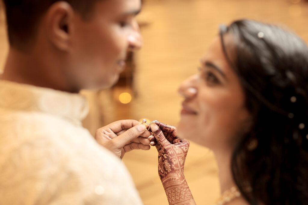 A close-up, shallow depth-of-field portrait focusing on a couple's hands as they show off their diamond engagement rings. The man wears a light embroidered kurta, and the woman, adorned with intricate henna (mehendi) on her hand, smiles lovingly at him while their faces are softly blurred in the foreground and background.