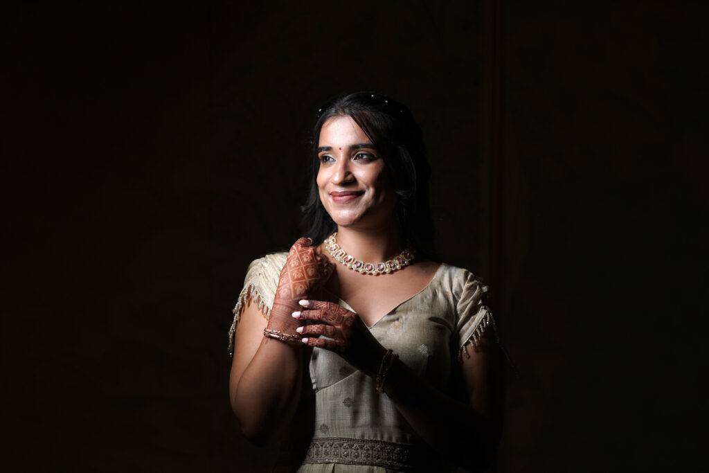 A dramatic, low-key indoor portrait of a bride smiling softly while bathed in bright, directional light against a deep, dark background. She wears an embroidered ivory outfit, a beaded necklace, and intricate mehendi on her hands. Part of a photographer's silhouette is visible in the foreground left shadow.