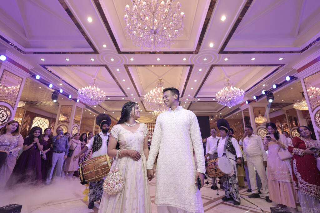 A wide-angle indoor photo of a couple walking hand-in-hand through a grand, crystal-chandeliered banquet hall. They are surrounded by family, guests, and traditional dhol players drumming along. The couple wears matching ivory and cream traditional attire, smiling lovingly at each other amidst low-lying fog effects.
