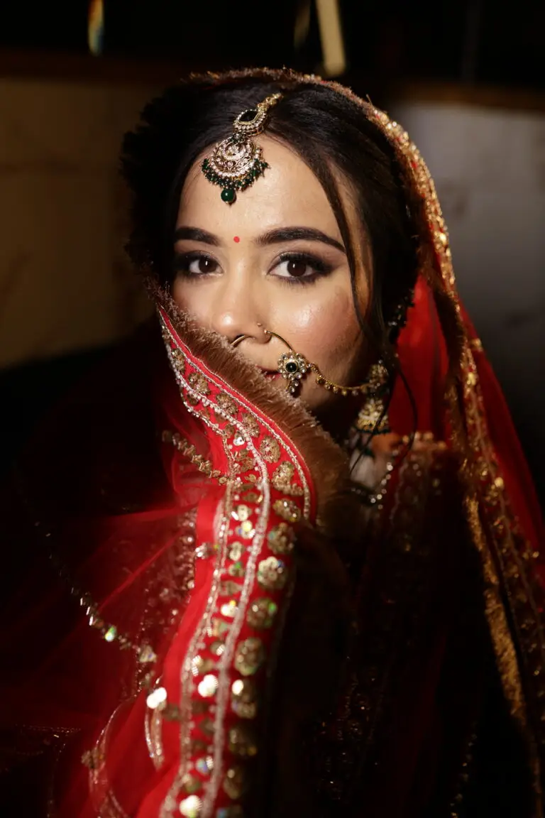 A dramatic close-up portrait of an Indian bride looking directly at the camera. She is holding a sheer red dupatta with heavy gold sequin embroidery over the lower half of her face, highlighting her expressive eyes, small red bindi, and traditional kundan jewelry featuring green stones.