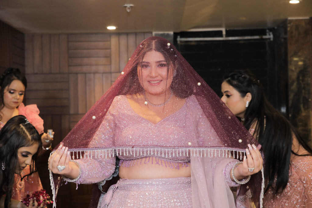An indoor portrait of a smiling bride holding up the edge of a deep maroon, pearl-adorned sheer veil draped over her head. She wears a shimmering lilac-pink embroidered lehenga, a delicate necklace, and bracelets, surrounded by bridesmaids getting ready in the background.