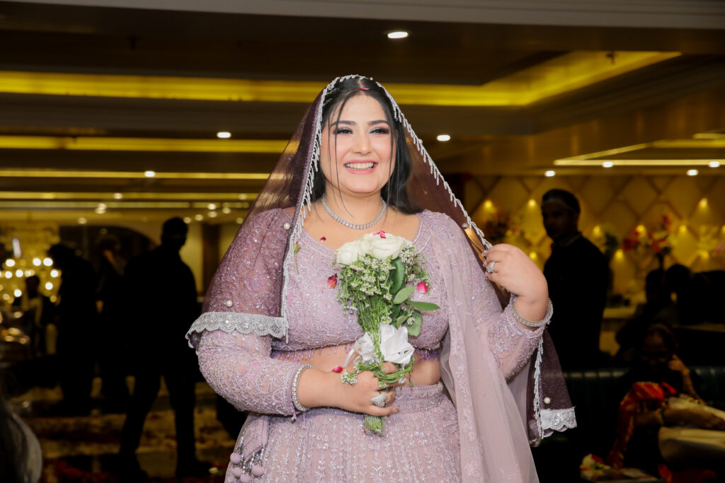 An indoor portrait of a smiling bride holding a bouquet of white roses and greenery. She wears an embroidered lilac-pink lehenga, a sparkling diamond necklace, bracelets, and a matching sheer veil draped over her head with beaded detailing.