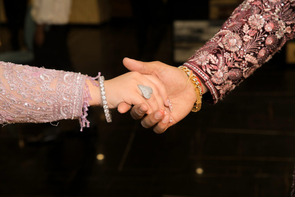 A close-up, macro shot of a couple holding hands gently. The person on the left wears a light pink embellished sleeve, a sparkling diamond bracelet, and a large heart-shaped diamond ring. The person on the right wears a heavily embroidered maroon and floral sleeve with a gold bracelet.