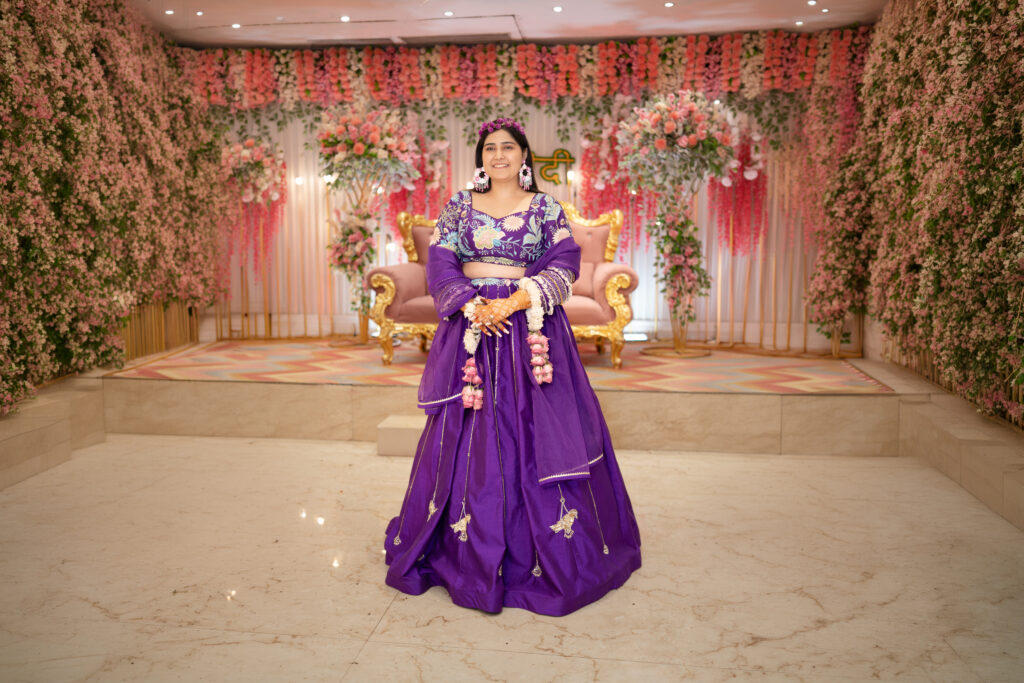 An indoor full-length portrait of a smiling woman standing on a polished floor, wearing a rich royal-purple lehenga with delicate embroidery and hanging tassels. She wears floral jewelry and a floral headband, set against a stage beautifully decorated with pink and white flowers and a pink velvet couch.