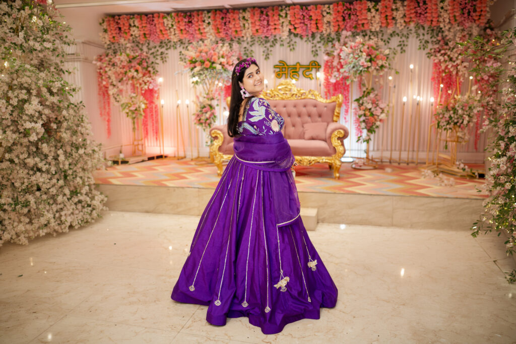 An indoor portrait of a smiling woman looking back over her shoulder while wearing a rich royal-purple lehenga with intricate silver line embellishments and bird motifs. She wears a floral headband and matching earrings, set against a stage decorated with pink and white flowers and a pink couch with the Hindi word "Mehendi" in lights.