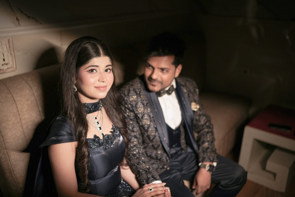 An indoor portrait of a couple seated together on a plush upholstered couch in a warmly lit, moody room. The woman looks toward the camera wearing a dark blue embellished gown and layered diamond necklace, while the man sits beside her in a patterned tuxedo jacket and bow tie, looking lovingly at her.