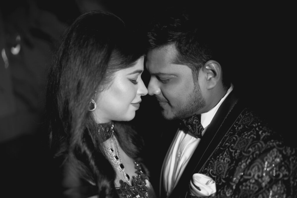 An intimate, close-up black-and-white portrait of a couple standing forehead-to-forehead with their eyes closed. The woman wears traditional jewelry and an embellished gown, and the man wears a patterned tuxedo jacket with a bow tie, set against a dark background.
