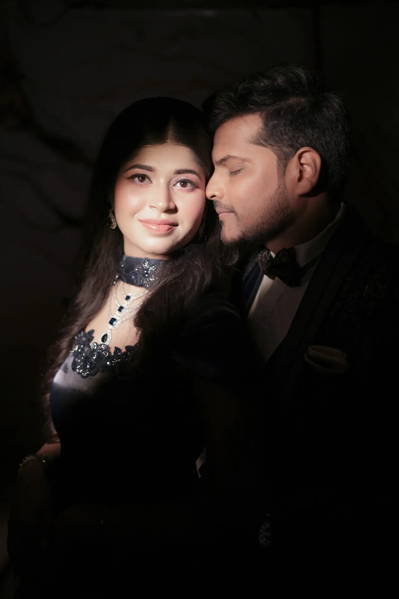 A dramatic, low-light close-up portrait of an engaged couple. A directional spotlight illuminates the woman's face, dark blue embellished gown, and layered necklace, while the man, wearing a dark patterned tuxedo, rests his face affectionately against hers in the shadows.