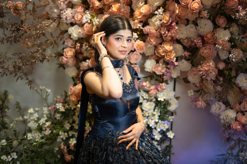 An indoor portrait of a woman posing against a lush wall of pastel pink, peach, and white roses. She wears a dark blue embellished gown with delicate beadwork and a matching dupatta, accessorized with sparkling bracelets and jewelry while gently touching her hair.