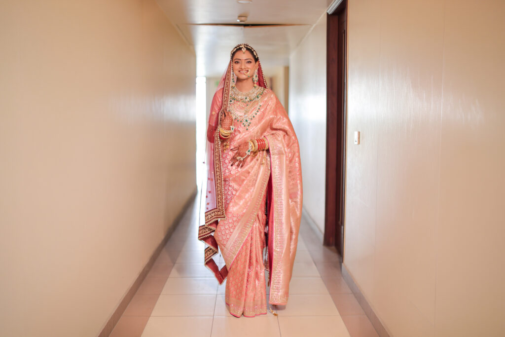 A full-length indoor portrait of a smiling bride standing in a brightly lit hallway, wearing a peach-pink traditional silk saree, a matching red-bordered veil, and heavy layered bridal jewelry including a choker necklace and traditional bangles.