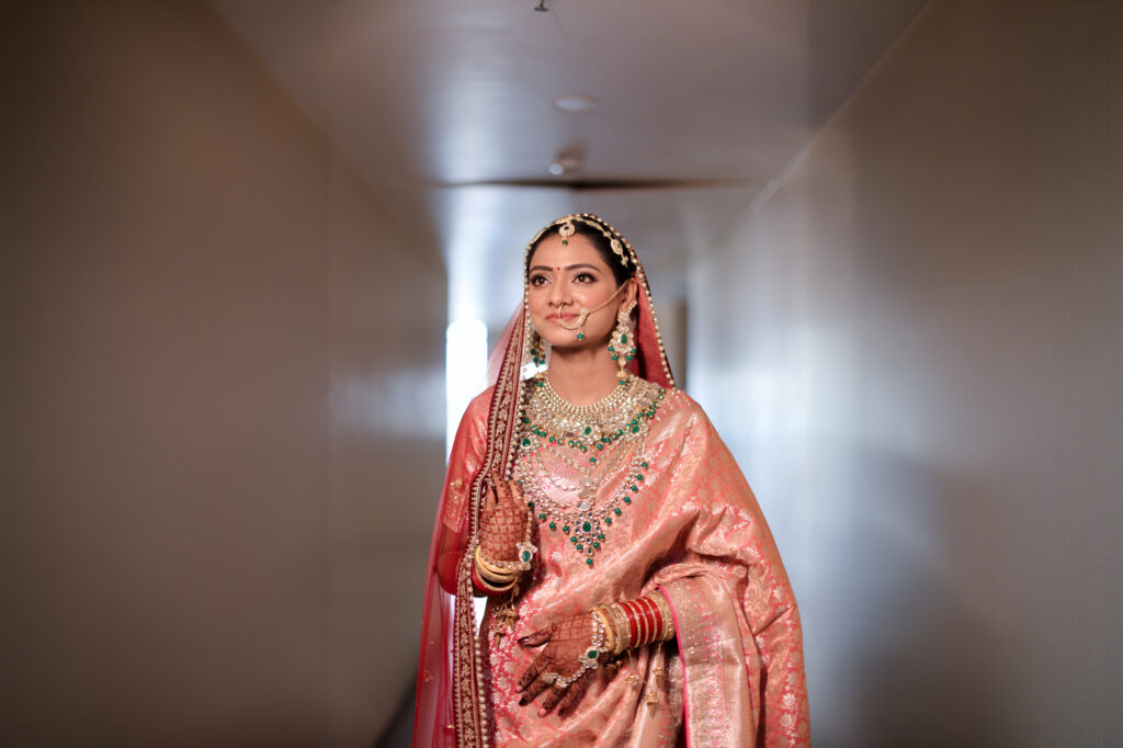 A medium indoor portrait of a bride posing gracefully in a corridor, wearing a peach-pink traditional silk saree, a red-bordered veil, and layered statement jewelry with green gemstone accents.