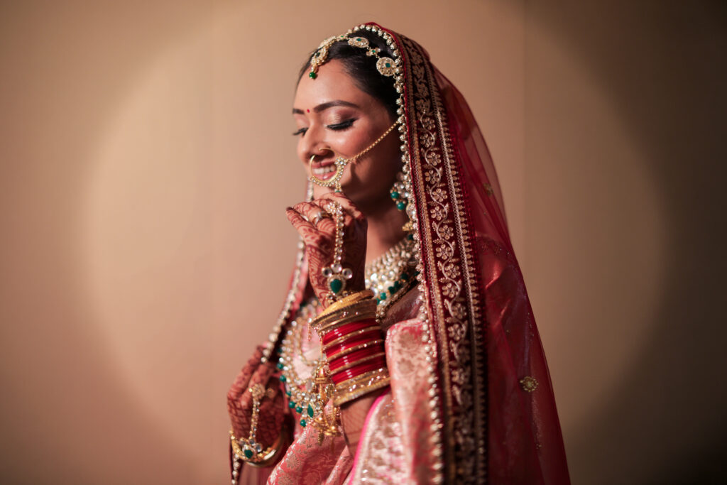 A close-up indoor portrait of a smiling bride wearing a sheer red embroidered veil and traditional wedding attire. She is adorned with classic statement jewelry, including a nath (nose ring), maang tikka, choker necklace, and red bridal bangles, set against a warm spotlighted background.