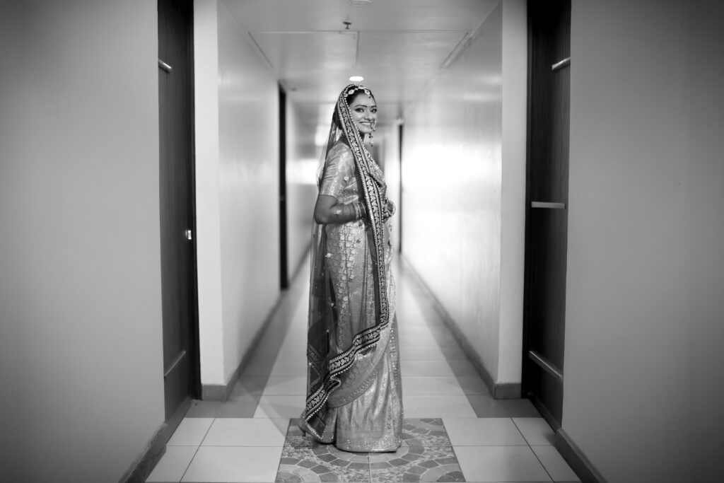 A black-and-white full-length portrait of a smiling bride standing in a modern indoor corridor, looking back over her shoulder. She wears a traditional saree and veil, beautifully captured in monochrome tones.