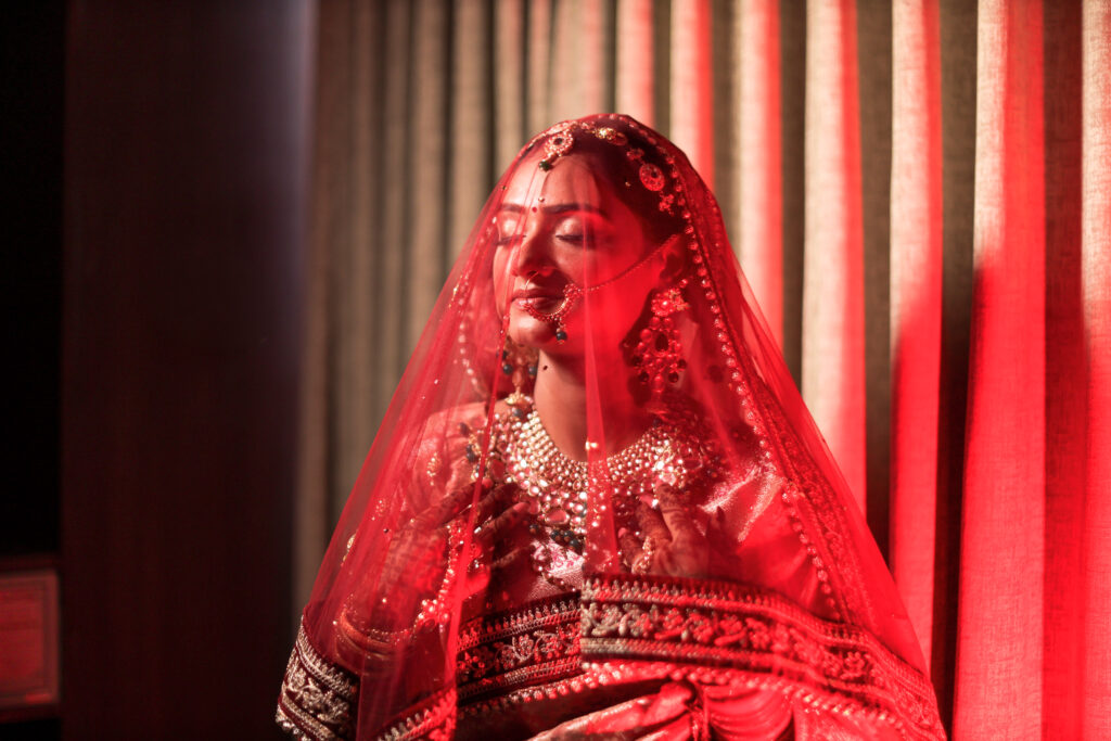 An indoor portrait of a bride with her eyes closed peacefully, draped in a sheer, vibrant red bridal veil. She is adorned with traditional statement jewelry, including a heavy necklace, earrings, a maang tikka, and a nose ring, set against a warm red-toned curtain background.