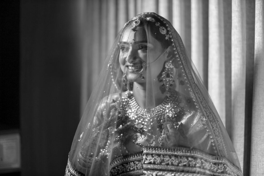 A black-and-white indoor close-up portrait of a smiling bride wearing a sheer, embellished veil draped over her head. She is adorned with heavy traditional jewelry, including a statement necklace, earrings, and a nose ring, set against a curtain background.