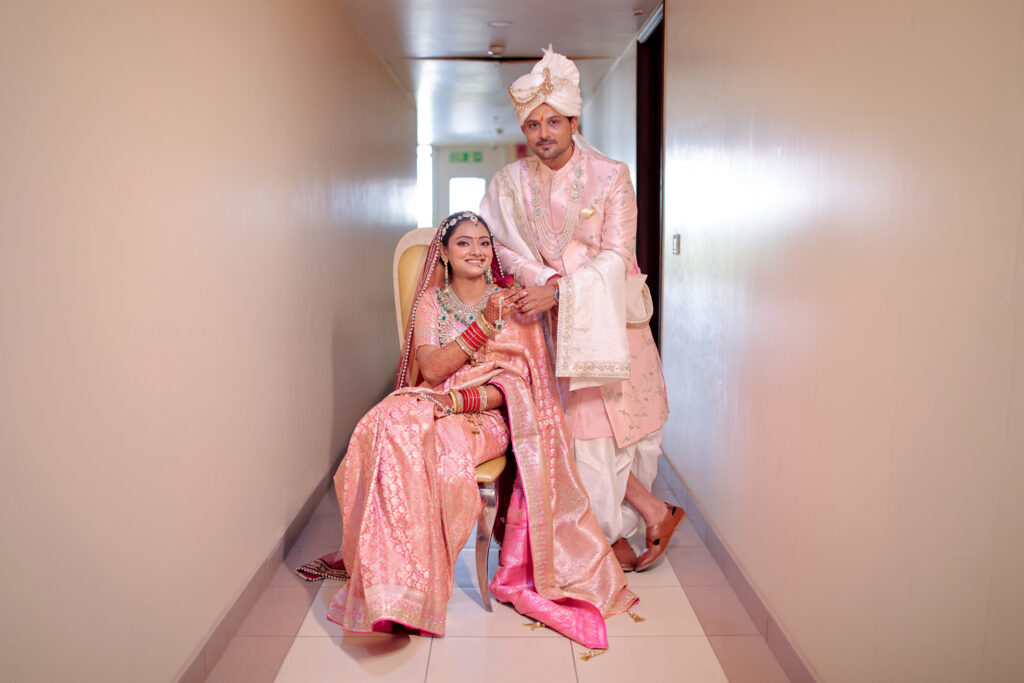 An indoor portrait of a newlywed couple posing in a brightly lit hallway. The bride is seated on a chair, wearing a shimmering pastel-pink traditional silk saree and matching veil, while the groom stands beside her wearing a coordinated light pink sherwani and turban.