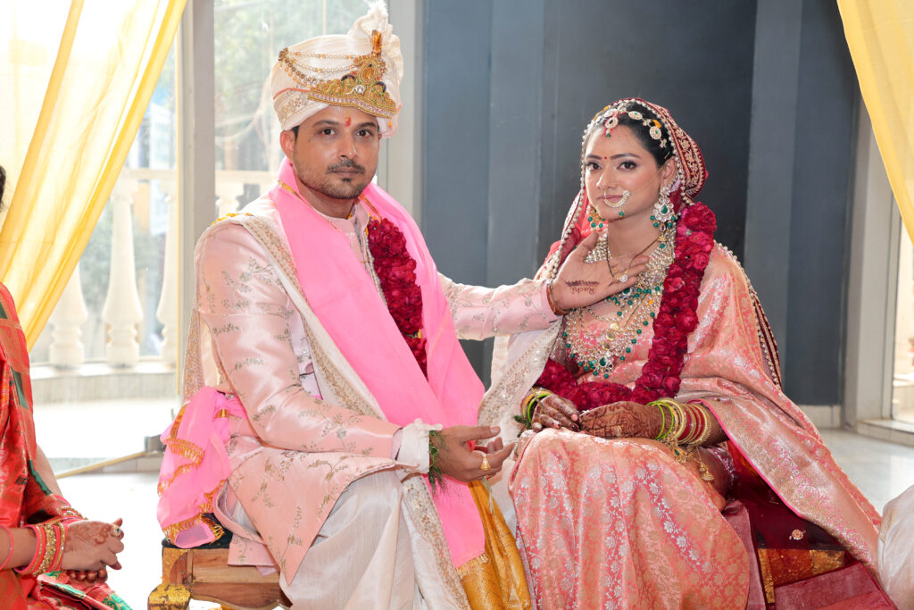 An indoor portrait of a newlywed couple seated together during a traditional ceremony. The groom wears a light pink sherwani with embroidery, a matching turban, a bright pink stole, and a red floral garland. The bride wears a traditional pink-and-gold outfit, a red veil, a heavy statement necklace, and a red floral garland, with her hand gently touching her mangalsutra.