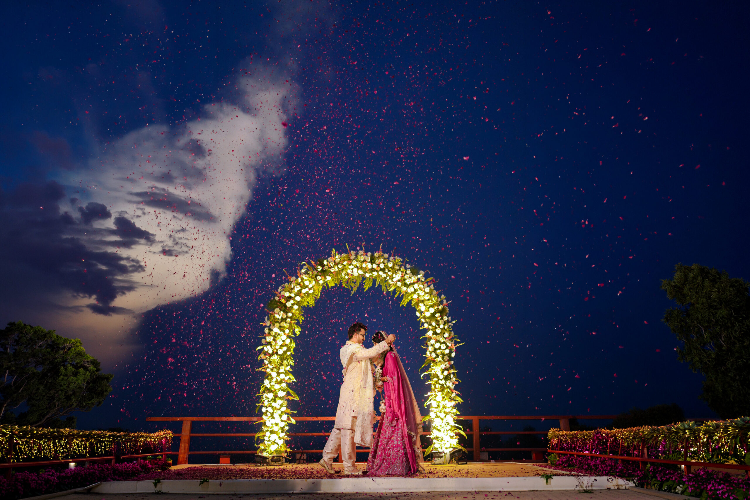 A wide, cinematic outdoor portrait of a newlywed couple standing beneath a glowing, illuminated floral archway at dusk. A shower of red and pink flower petals falls through the night sky around them, with dramatic clouds and lights framing the scene.