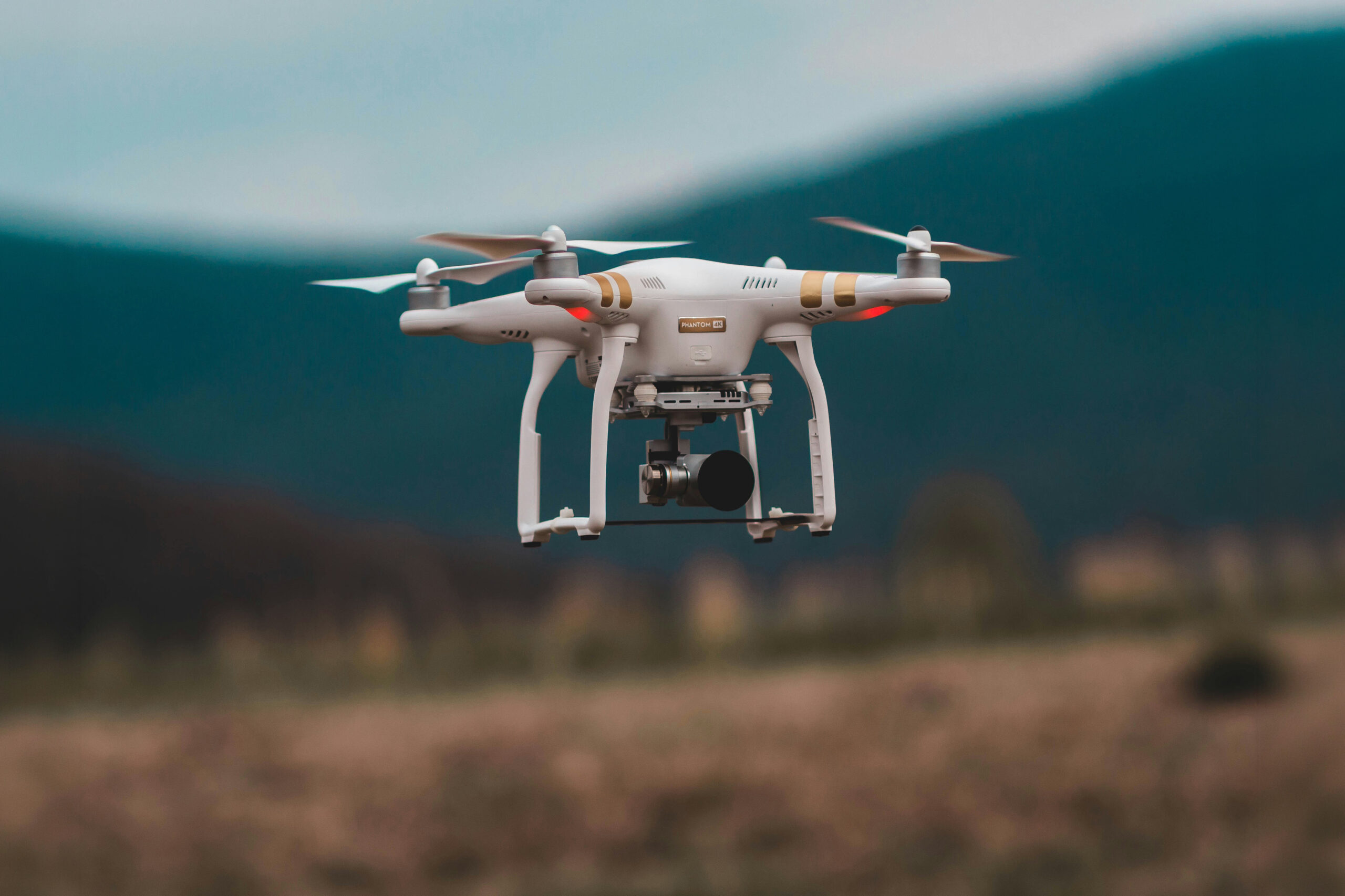 A close-up shot of a white professional quadcopter drone with an integrated camera hovering mid-air against a soft-focus background of rolling hills and nature.